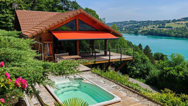 Una cabaña de madera con un toldo rojo, un jacuzzi y vistas a un lago y colinas.
