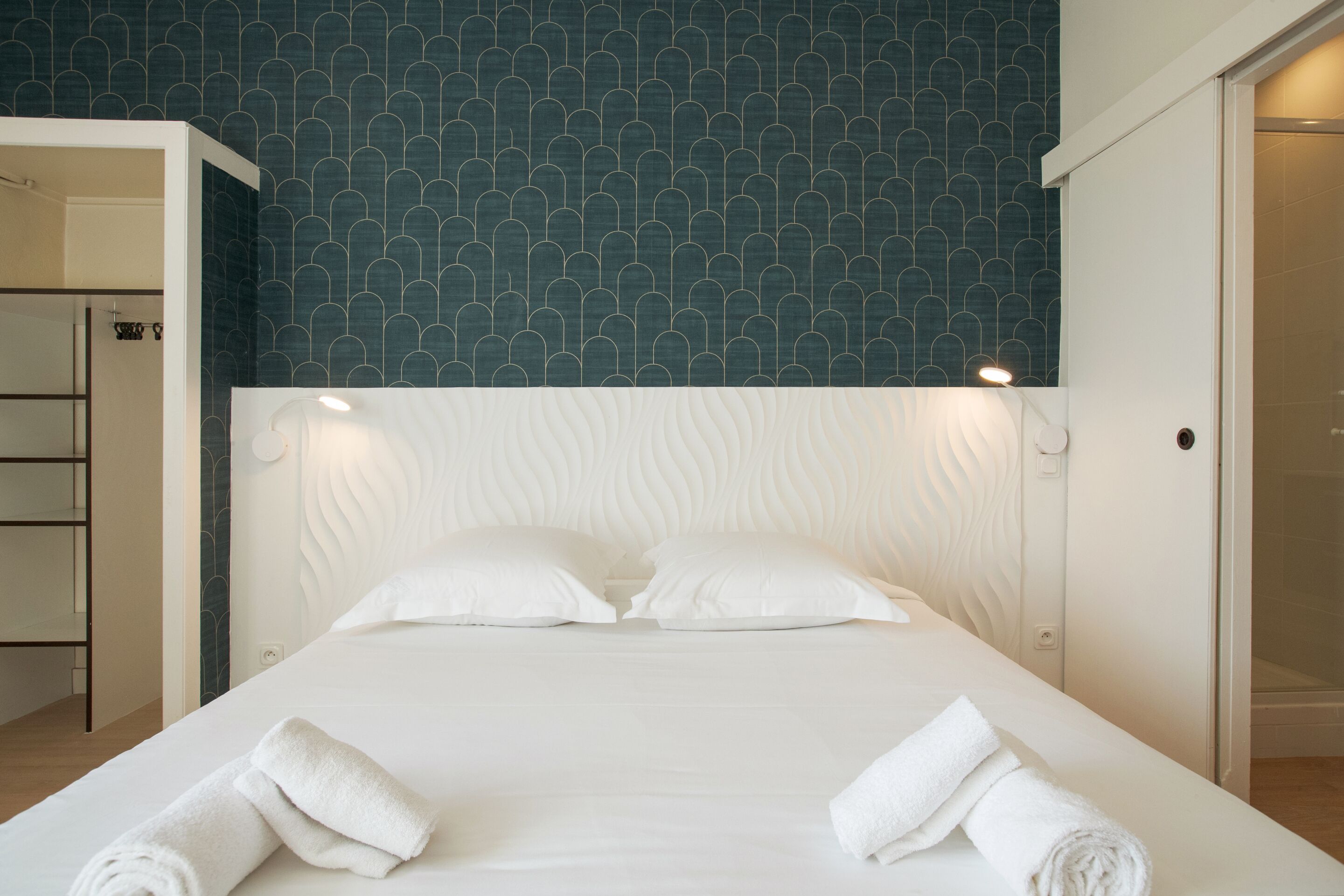 Een net opgemaakt bed met witte lakens en twee opgerolde handdoeken, een wit hoofdbord en een blauwe behang.