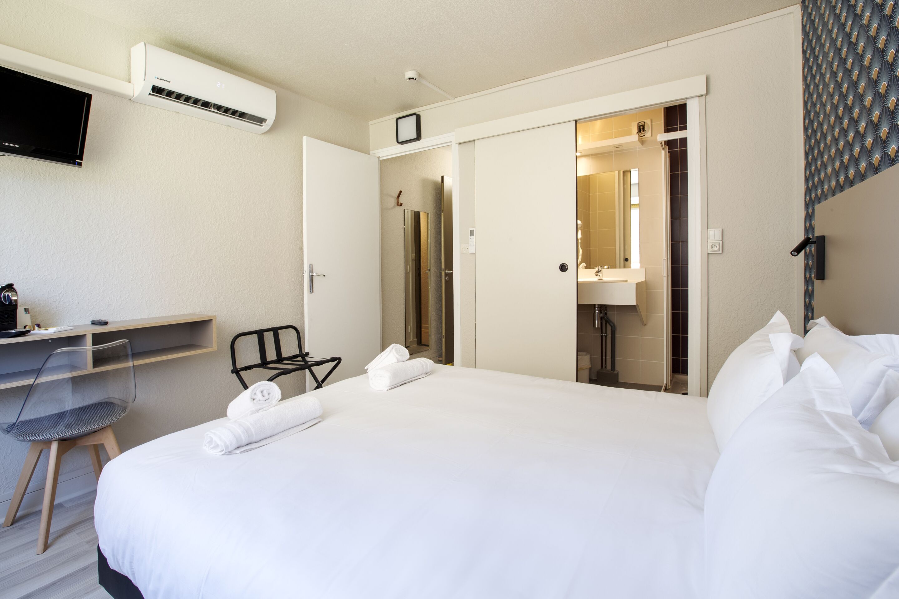 A hotel room with a bed, a desk with a chair, a television, and a bathroom visible through an open door.