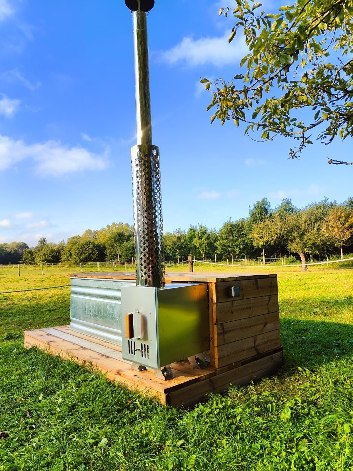 Ein tragbarer Holzofen mit Metallschornstein, auf einer hölzernen Plattform in einem grasbewachsen Feld platziert.