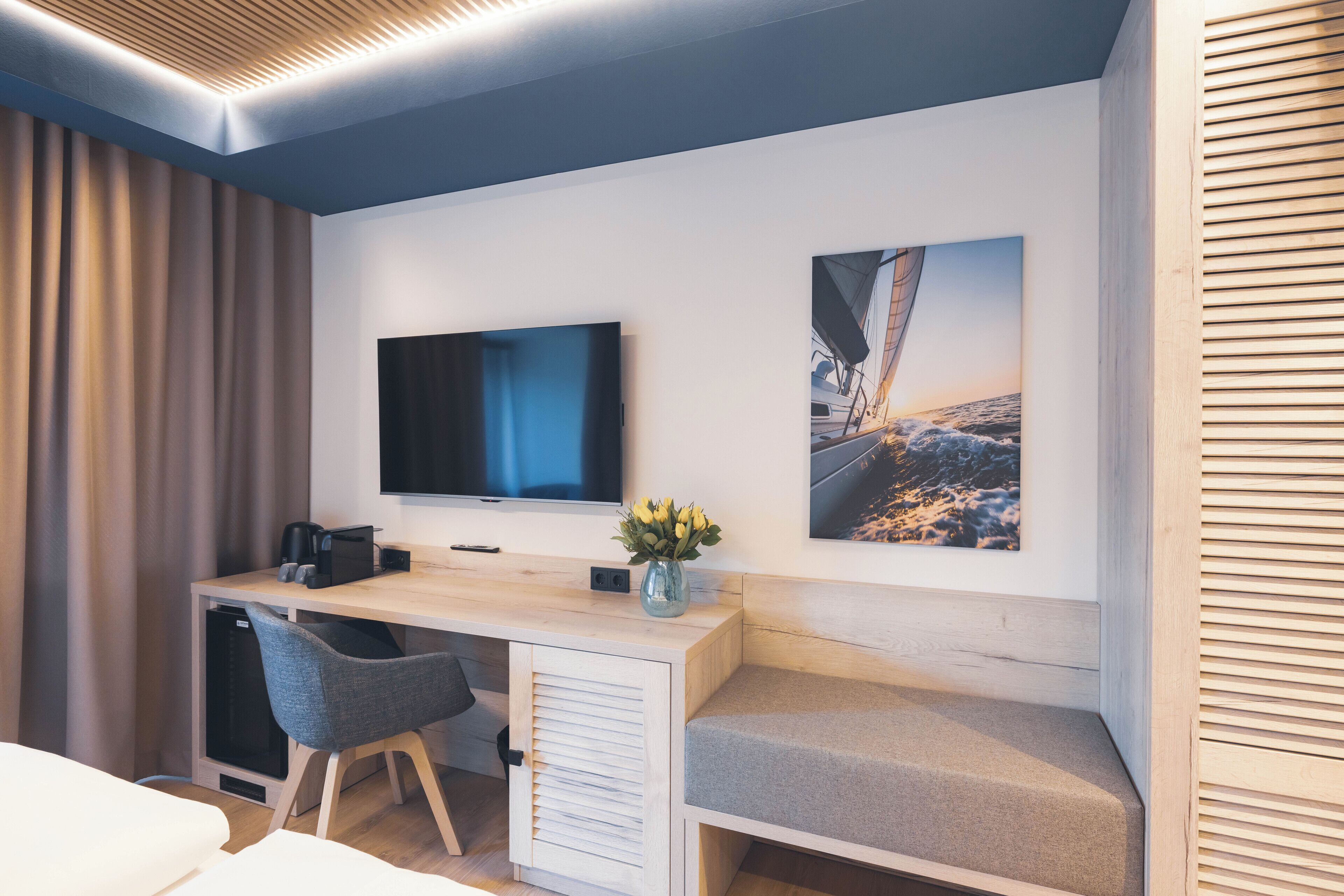 Ein modernes Hotelzimmer mit Schreibtisch aus Holz, einem Flachbildfernseher, einem bequemen Sessel, einer kleinen Sitzecke und einem großen Fenster mit Vorhängen.