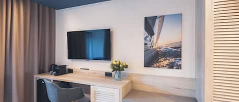 Una habitación de hotel moderna con un escritorio de madera, un televisor de pantalla plana, una silla cómoda, un pequeño rincón de descanso y un ventanal con cortinas.