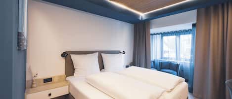 A modern hotel room with a large bed, a bedside table, a nightstand, and a window with curtains.