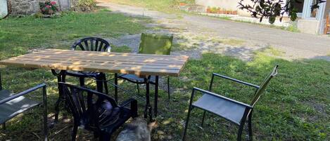 A rustic outdoor seating area with a wooden table and metal chairs, surrounded by a grassy lawn and a stone wall.