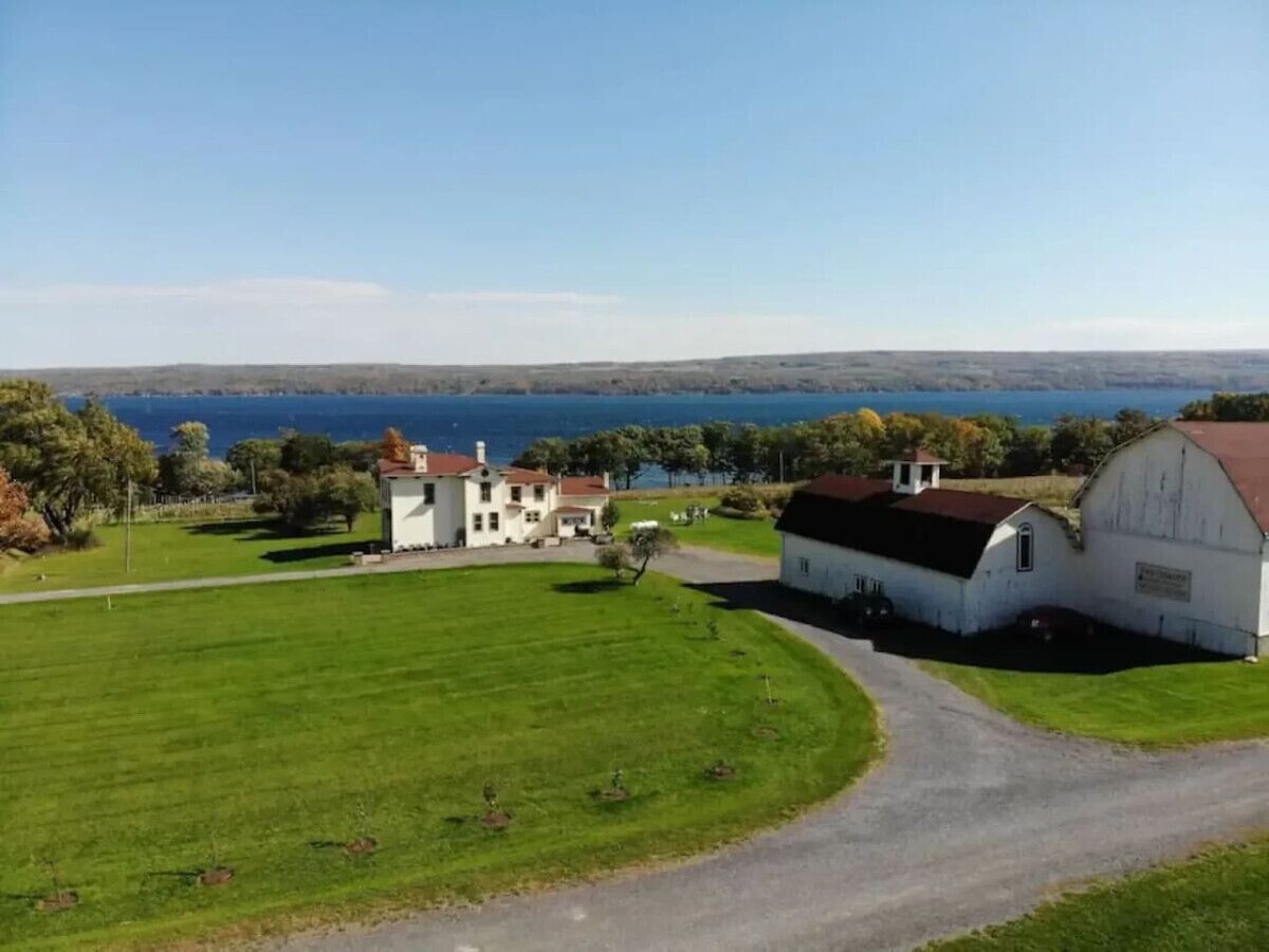 A large white barn and a white house with a red roof, surrounded by green fields and a view of the sea.