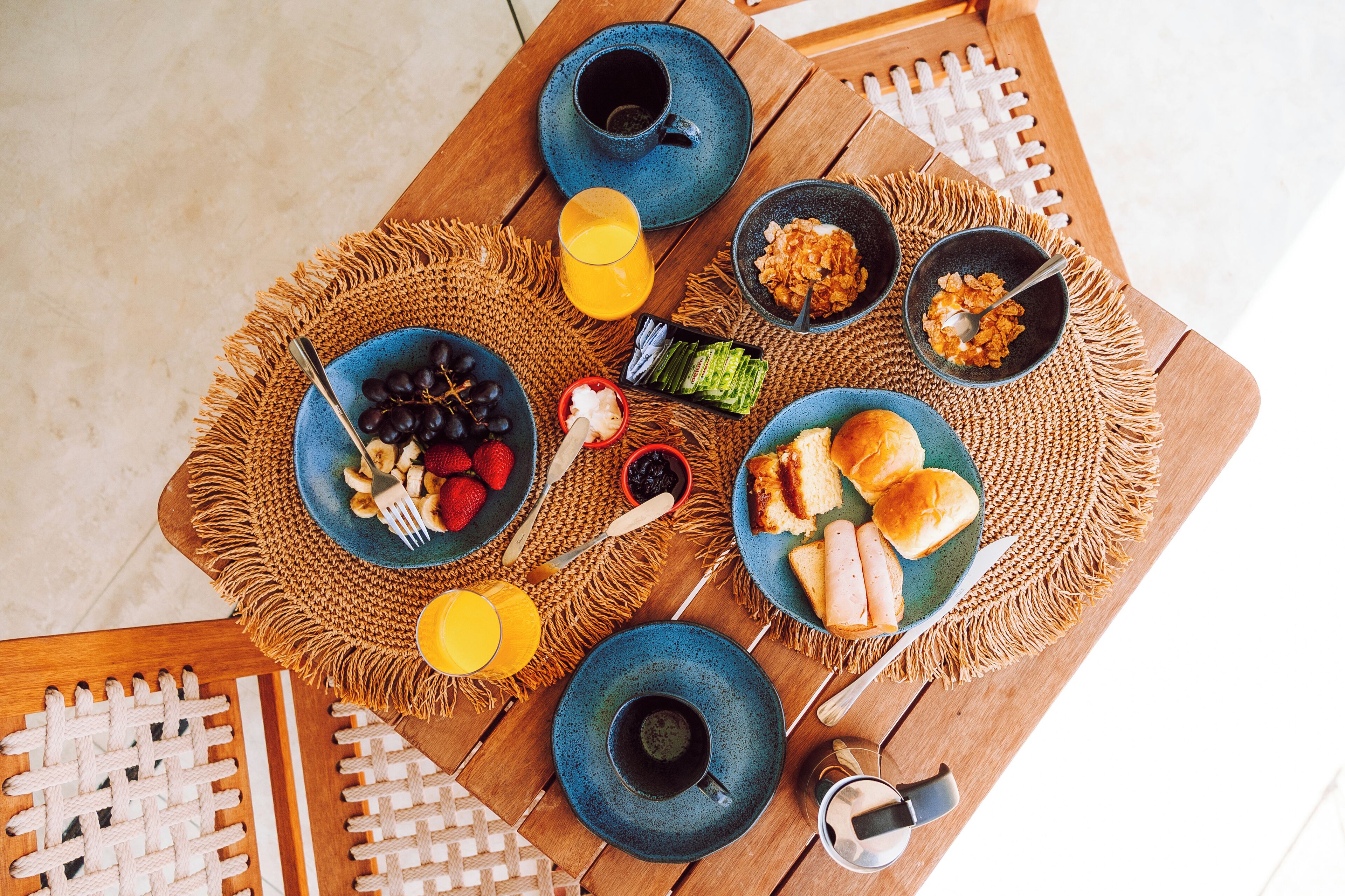 Een houten tafel met een geweven placemat, verschillende gerechten waaronder fruit, brood en een kom met ontbijtgranen, plus een kop koffie.