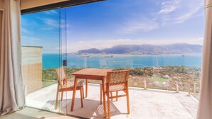 Un balcon avec une table et deux chaises, offrant une vue sur la côte, avec des montagnes et une étendue d’eau.