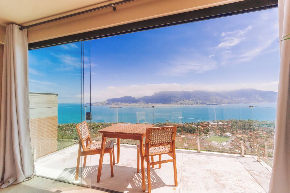 Un balcón con una mesa y dos sillas, con vistas a la costa, montañas y una masa de agua.