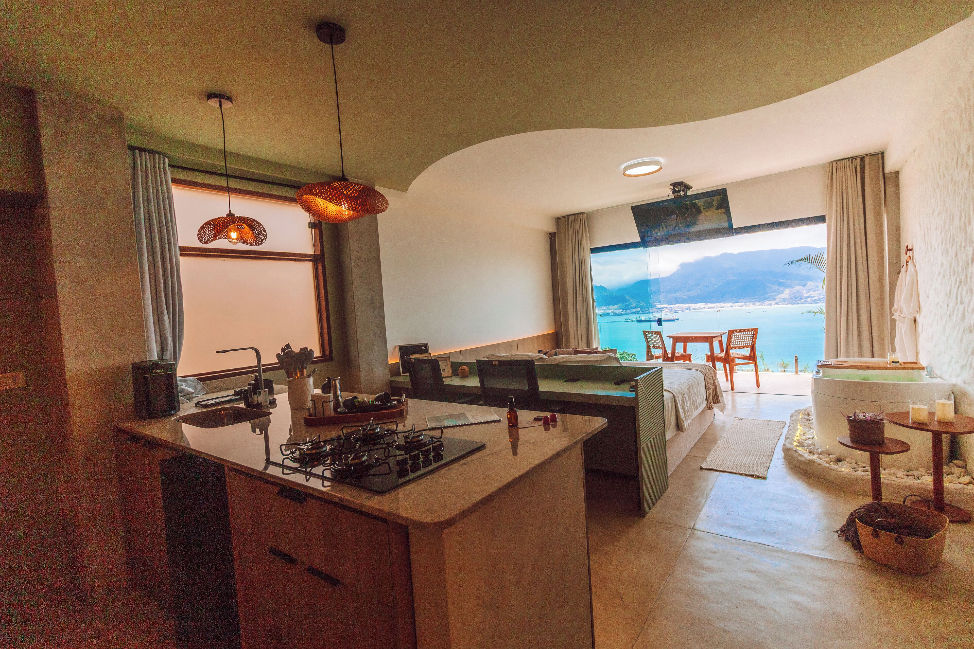 A modern kitchen with a central island, dining area with chairs, and a large window offering a view of the sea.
