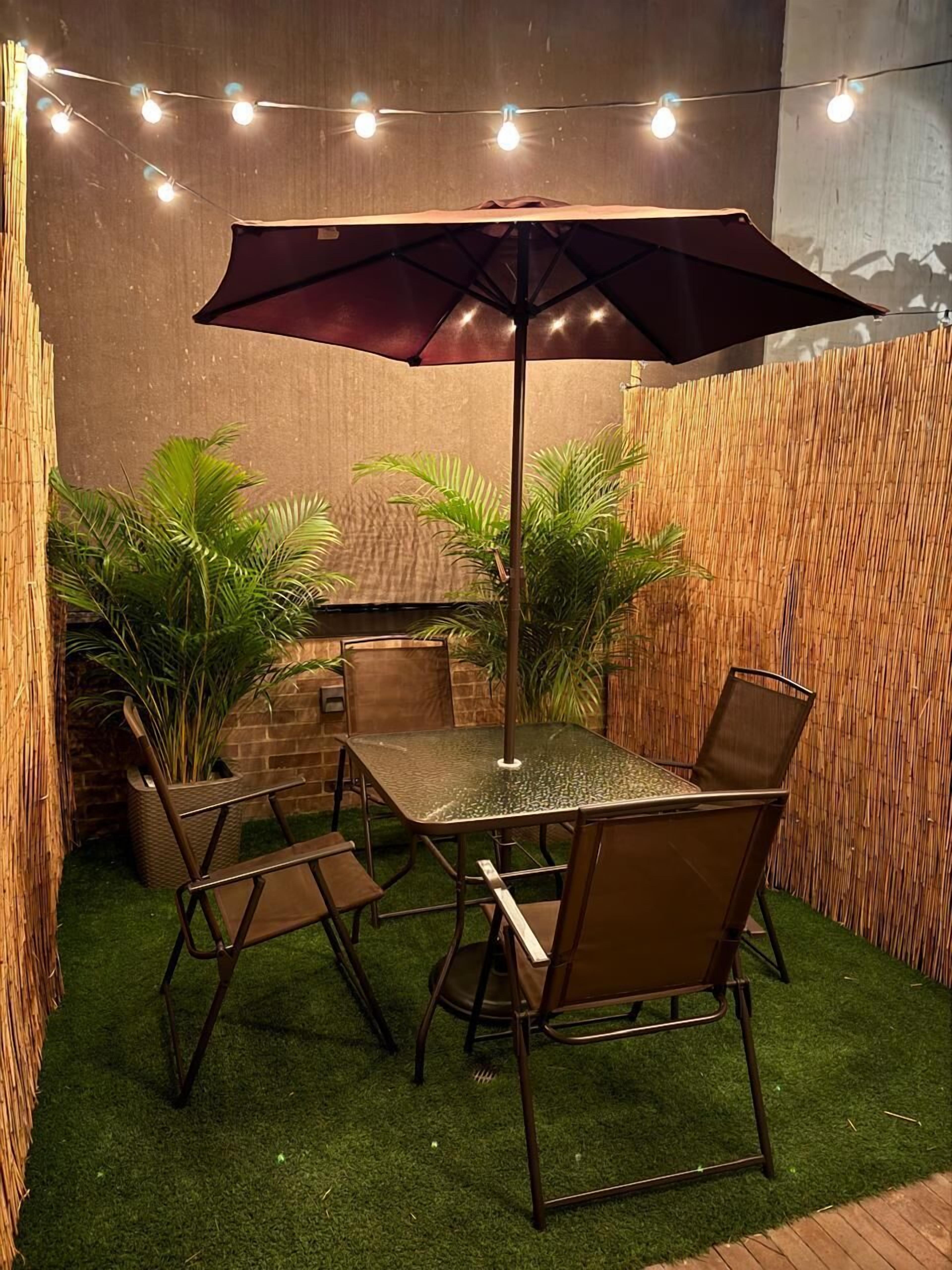 Ein Patio mit Tisch, Stühlen, Sonnenschirm, Lichterkette und Topfpflanzen.