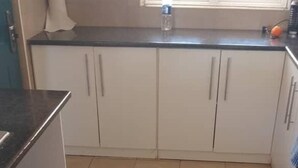 A small kitchen with white cabinets, a black countertop, and a window with blinds. - 3 Bed House - Sleeps 6 - Garden - Parking - Wifi (Lusaka)