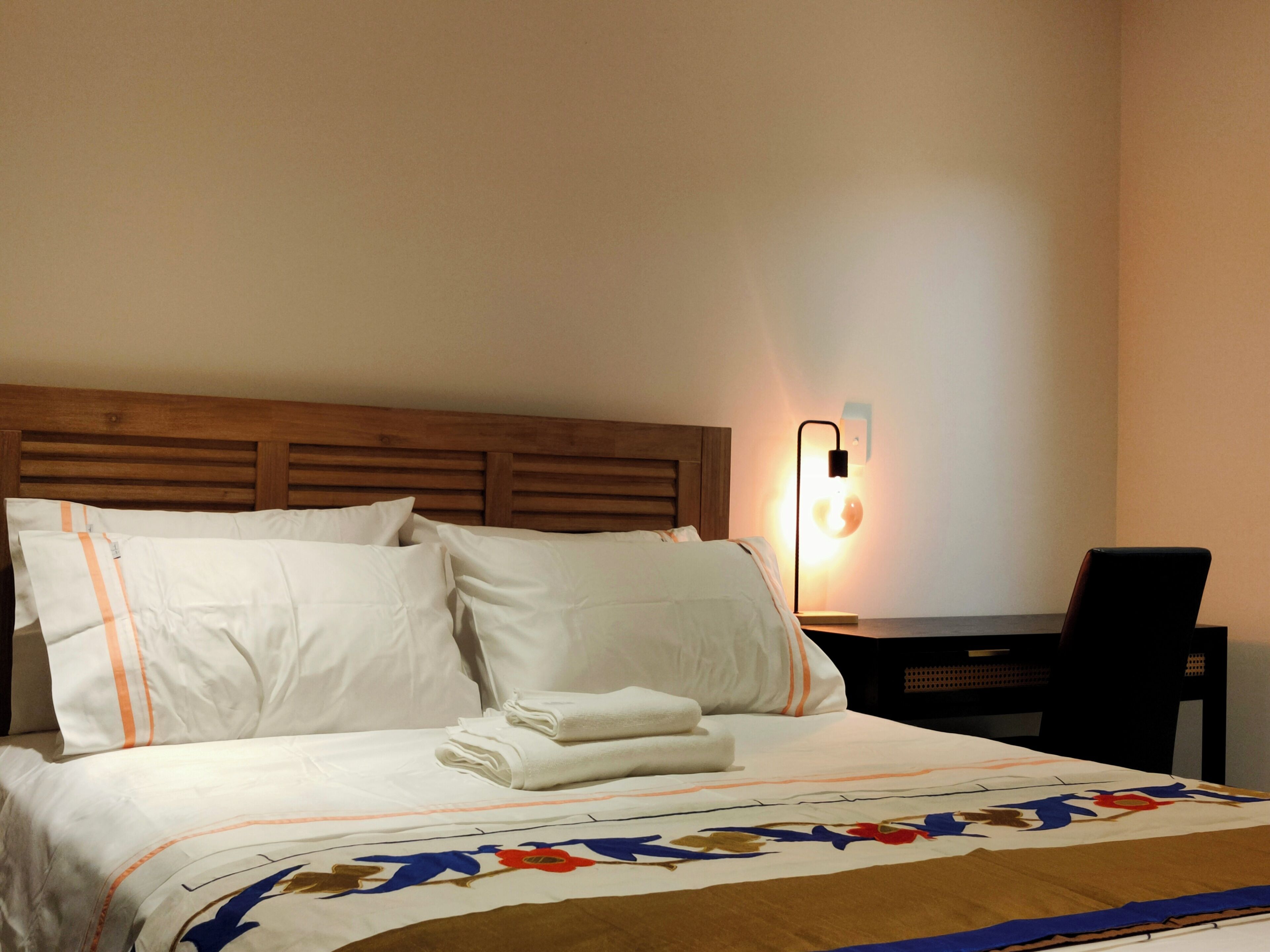 Ein ordentlich bezogenes Bett mit weißen Leinentüchern und einer Decke mit Blumenmuster, ein hölzernes Kopfende, ein Nachttisch mit Lampe und ein schwarzer Stuhl.