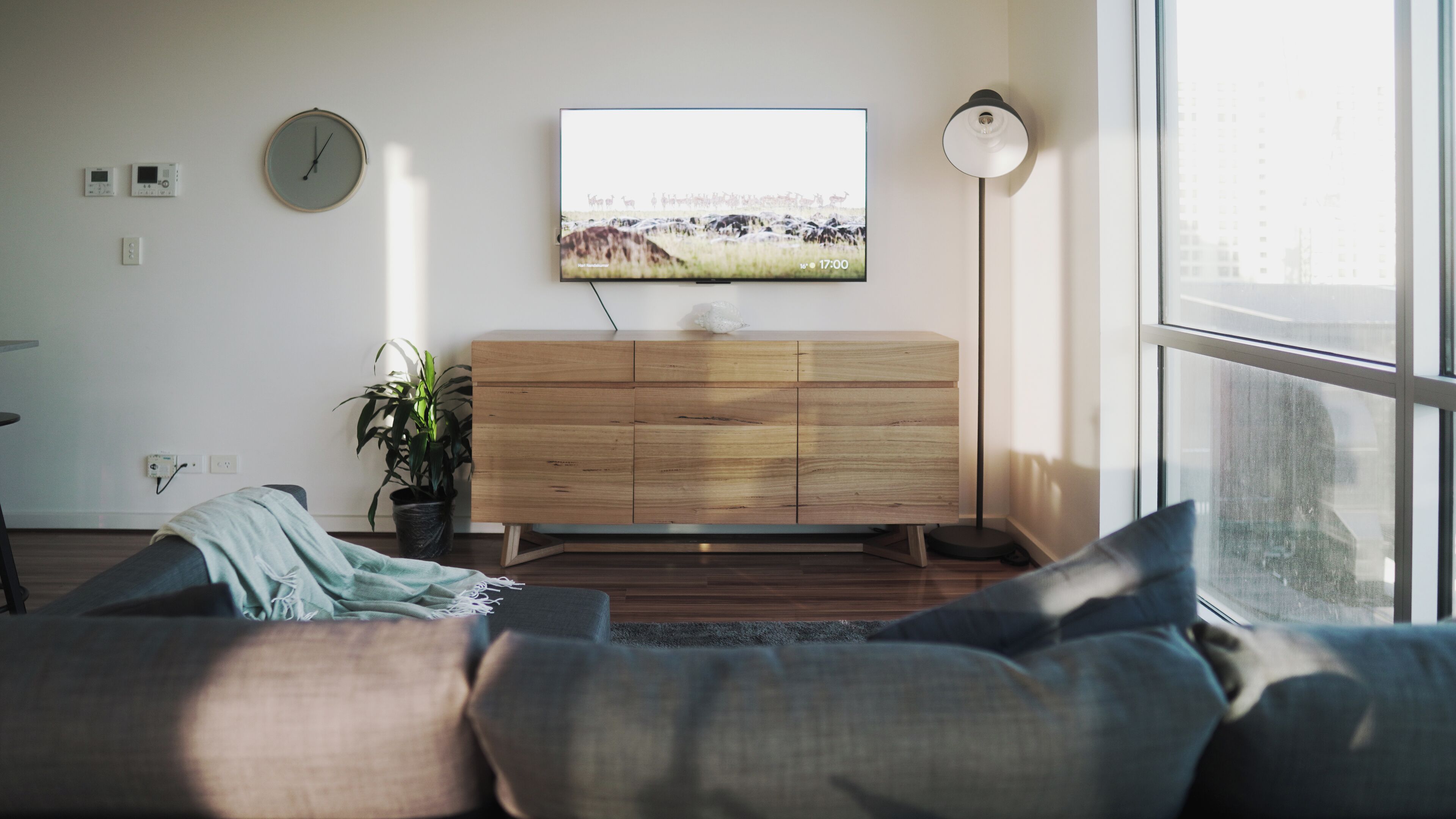 A modern living room with a wooden cabinet, a flat-screen TV, a floor lamp, and a large window.