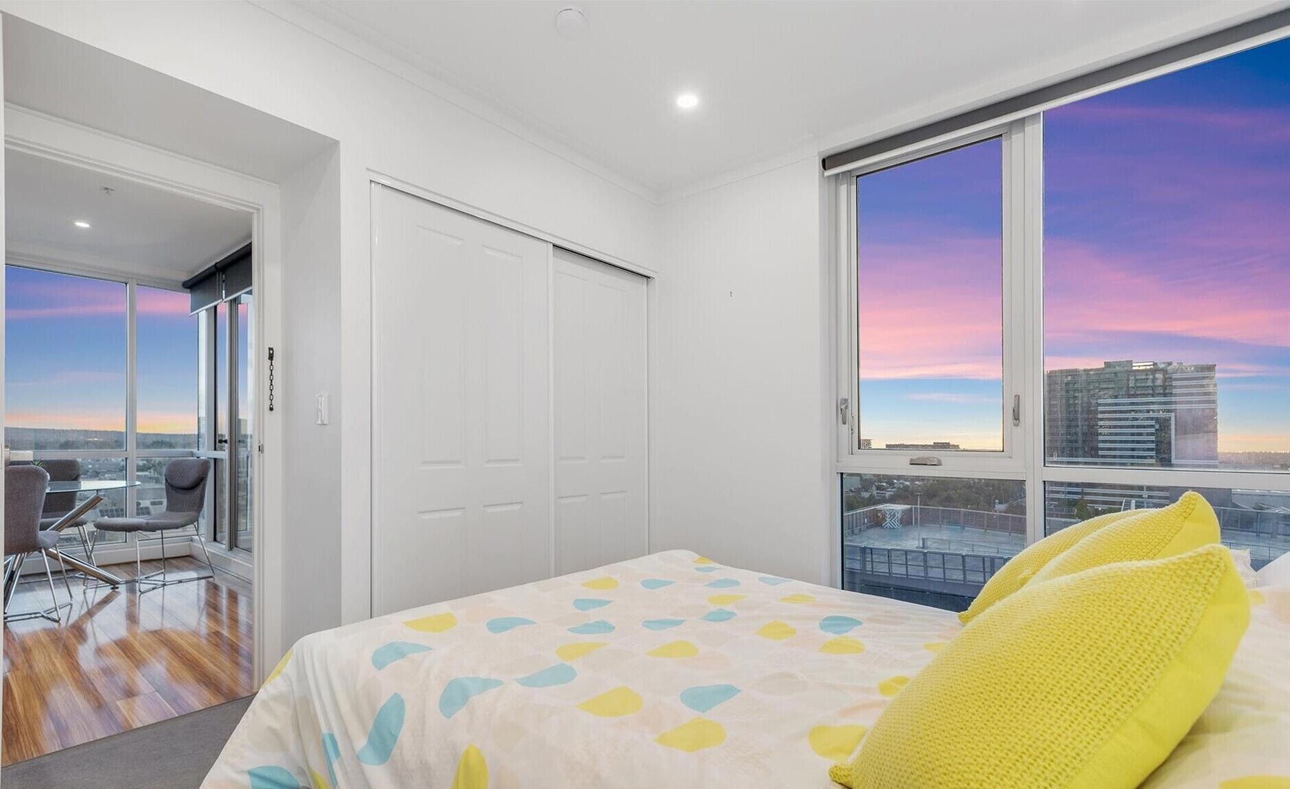 A modern bedroom with a large window, a bed with patterned bedding, and a view of the cityscape.