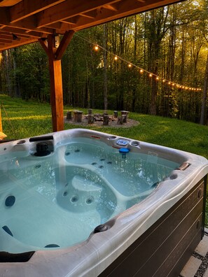 A hot tub in a covered outdoor area with a wooden deck, surrounded by a forest and string lights.