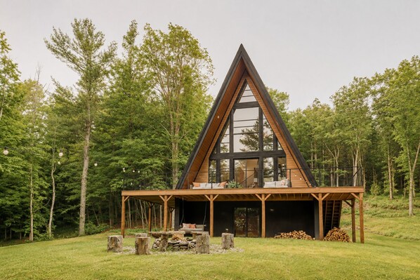 A modern A-frame cabin with a large glass window, wooden deck, and a fire pit.