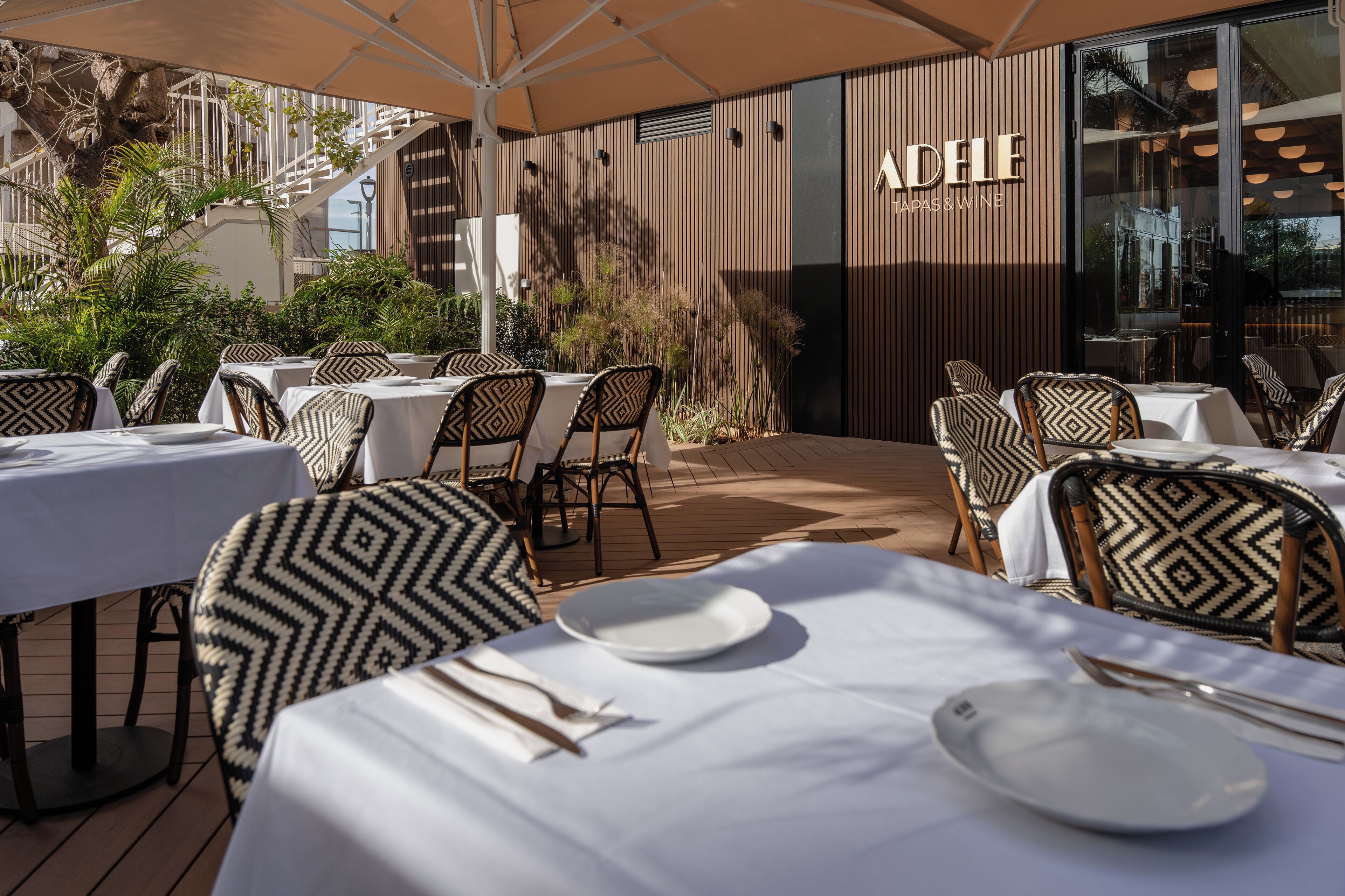Un área de comedor al aire libre con mesas y sillas, una escalera y un edificio con el rótulo 'ADELE JAPAN & WINE'.