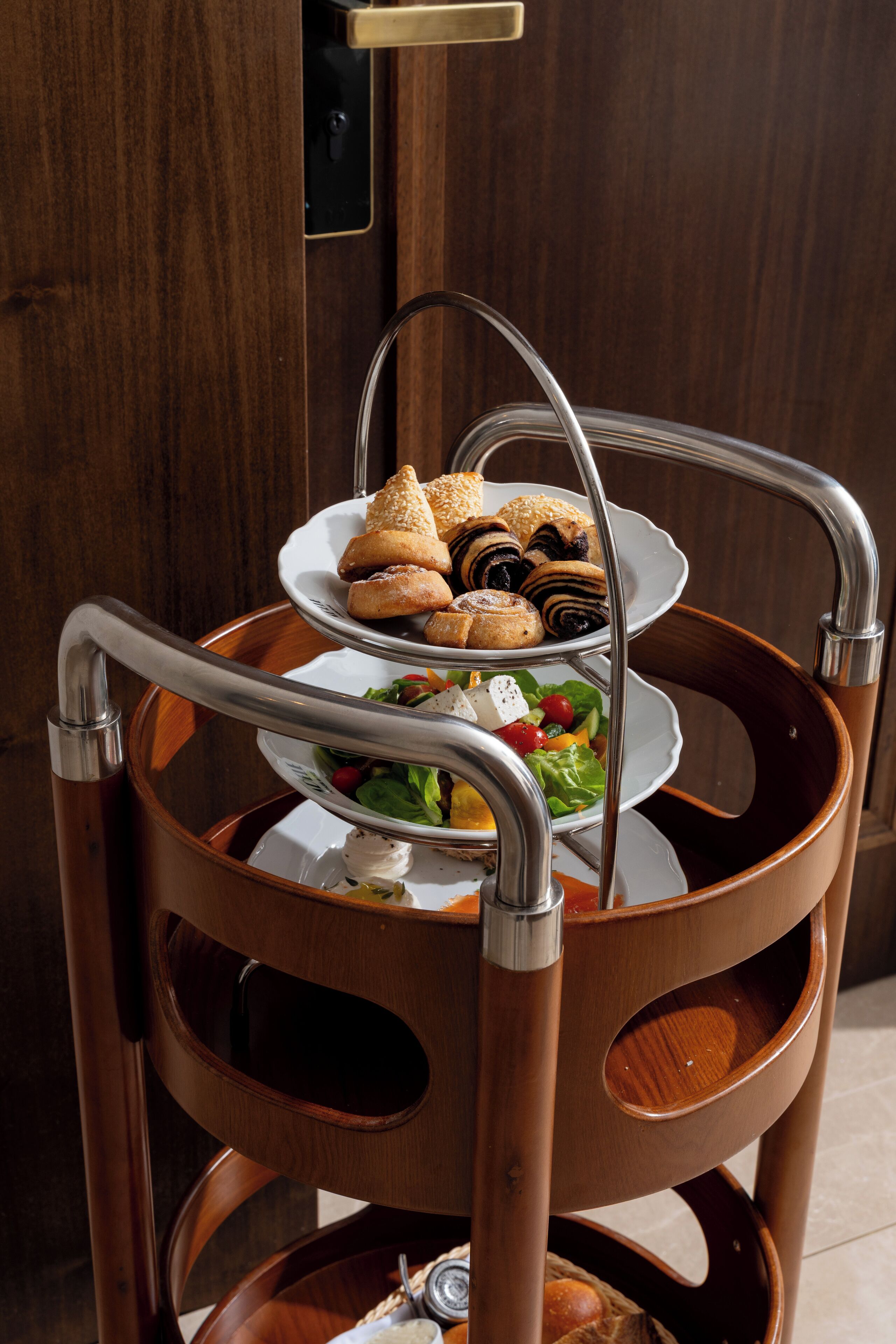 A three-tiered tray with pastries and a salad, placed on a wooden chair with a metal frame.
