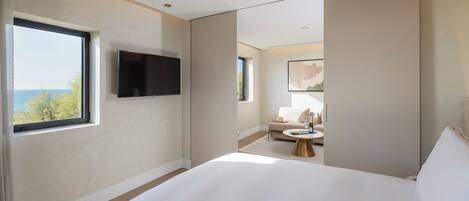 A modern hotel room with a large bed, a flat-screen TV mounted on the wall, a view of the sea through a large window, and a small seating area with a table and a bottle of wine.
