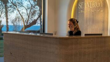 Una recepció amb un cartell que diu "SEANERGY Boutique & Spa Hotel", una finestra gran amb vista a arbres i il·luminació penjant.
