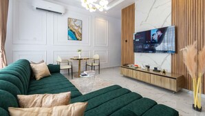 A modern living room with a green sectional sofa, a TV mounted on the wall, a wooden cabinet, and a marble-patterned wall. - Lux Lekki Home w PS5 Balcony 24 7 Power Security (Lekki)