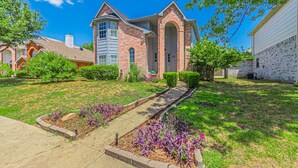 A residential house with a brick exterior, a front yard with a pathway, and a small garden area. - Lux 3-br Garage Gated Yard by The Star Frisco (Frisco)