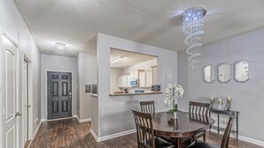 A dining area with a wooden table and chairs, a chandelier, and a kitchen in the background.