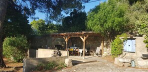 Ein rustikaler Außenbereich mit Steinsitzen, einer Holz-Pergola und einem Steinhaus mit blauer Tür.