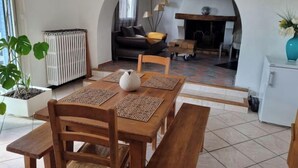 A cozy living room with a fireplace, a dining area with wooden furniture, and a large window. - Chambres D'hôtes La Magnanerie (Saint-Julien-de-Peyrolas)