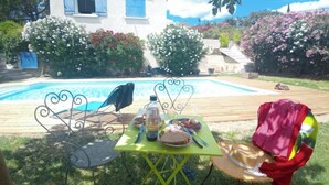 A patio with a table set for a meal, a bird on a chair, and a house in the background. - Chambres D'hôtes La Magnanerie (Saint-Julien-de-Peyrolas)