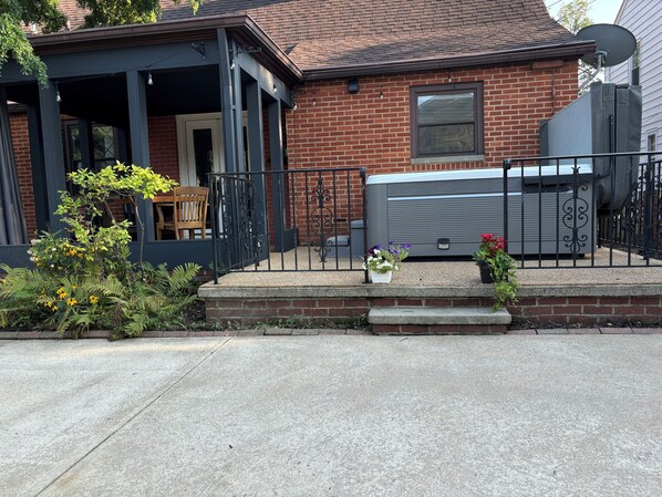 A brick house with a covered porch, a large air conditioning unit, and a small garden with flowers and shrubs.