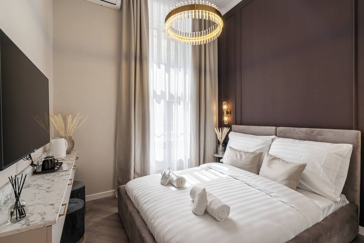 A modern hotel room with a large bed, a flat-screen TV, a desk with a coffee maker, and a large window with curtains.