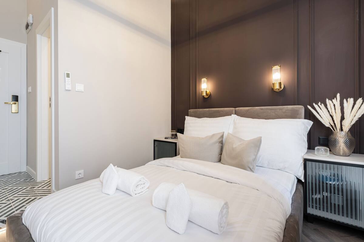 A modern hotel room with a large bed, two bedside tables, a nightstand, and a wall-mounted light fixture.