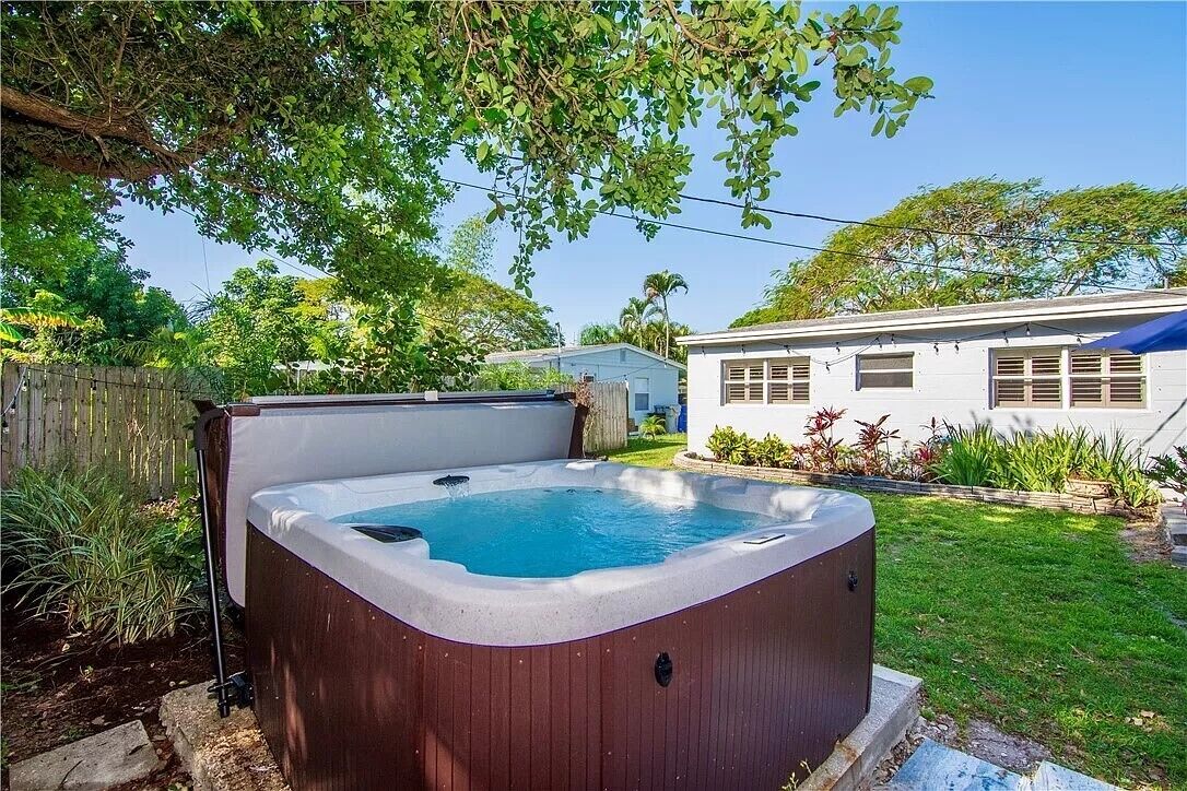 A hot tub in a backyard with a wooden fence, a white house, and a blue awning.