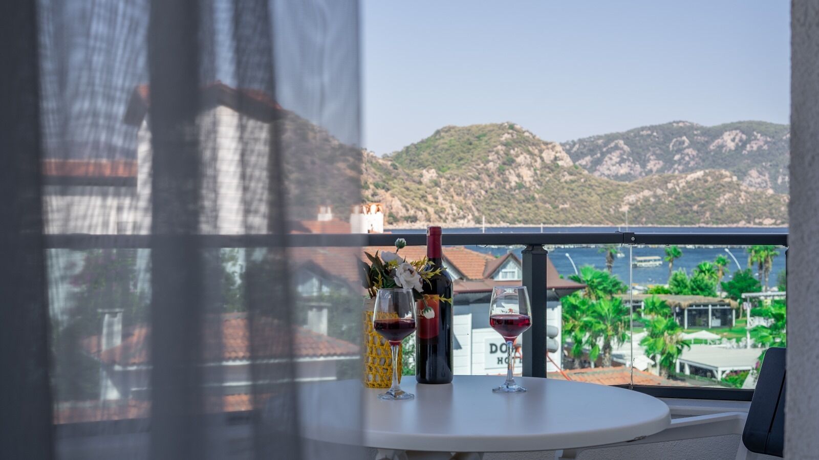 Un balcón con una mesa, una botella de vino y una copa de vino. Al fondo, un paisaje montañoso y un edificio con un cartel que dice "DOH HOTEL".