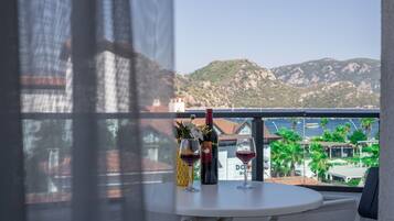 Un balcón con una mesa, una botella de vino y una copa de vino. Al fondo, un paisaje montañoso y un edificio con un cartel que dice "DOH HOTEL".