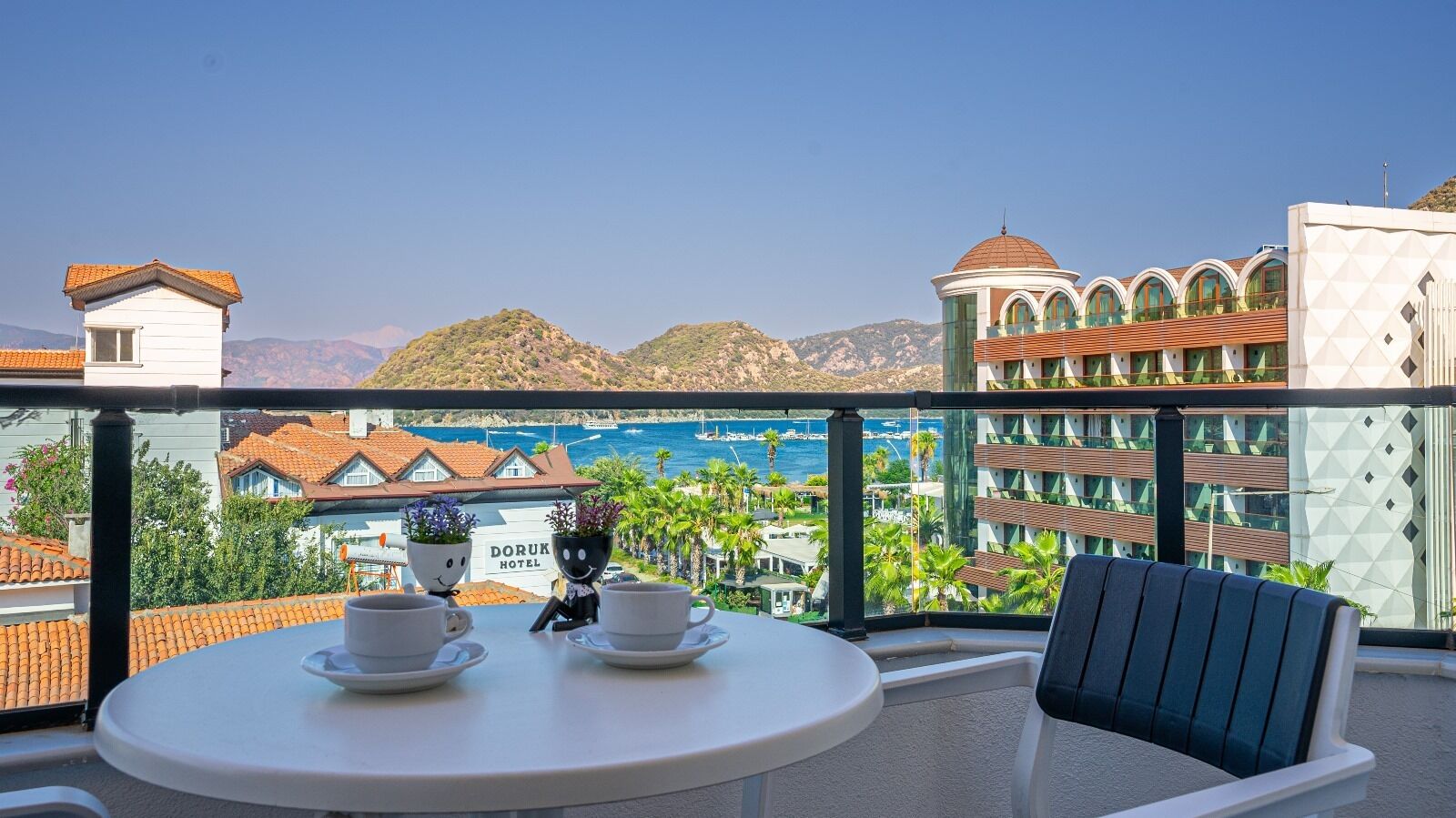 A balcony with a table and chairs, overlooking a coastal area with buildings and a clear sky.