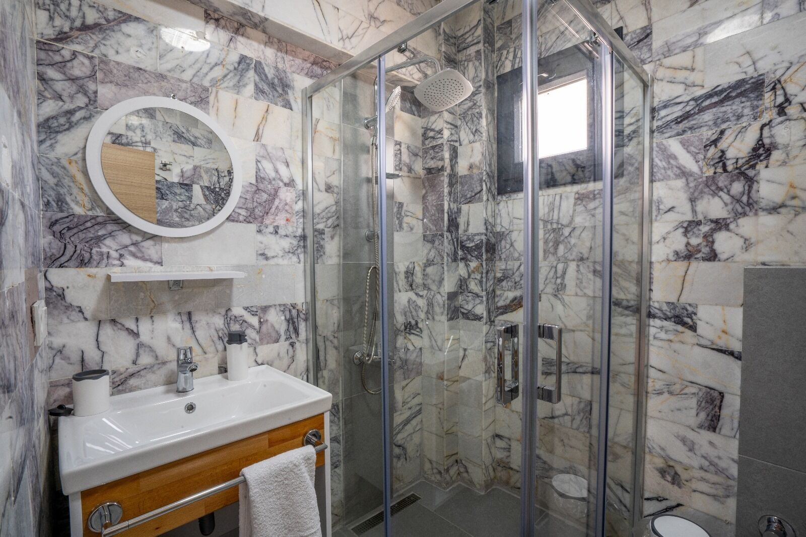 A bathroom with a marble wall, a glass-enclosed shower, a wooden vanity, and a round mirror.
