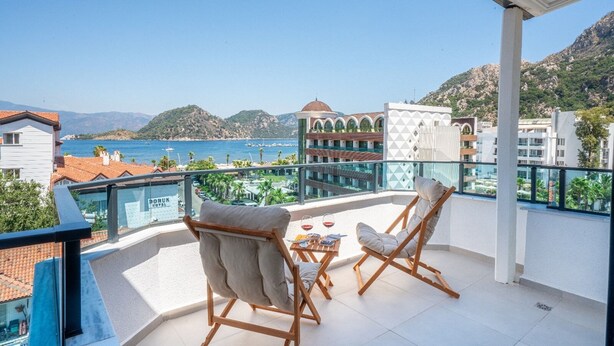 Un balcón con dos sillas y una mesita, con vistas a la costa, edificios y montañas.