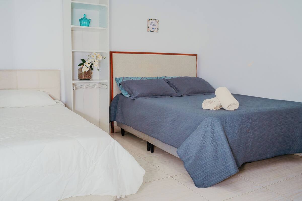 A bedroom with two beds, one with a white bedspread and the other with a blue bedspread. There is a shelf with decorative items and a framed picture on the wall.