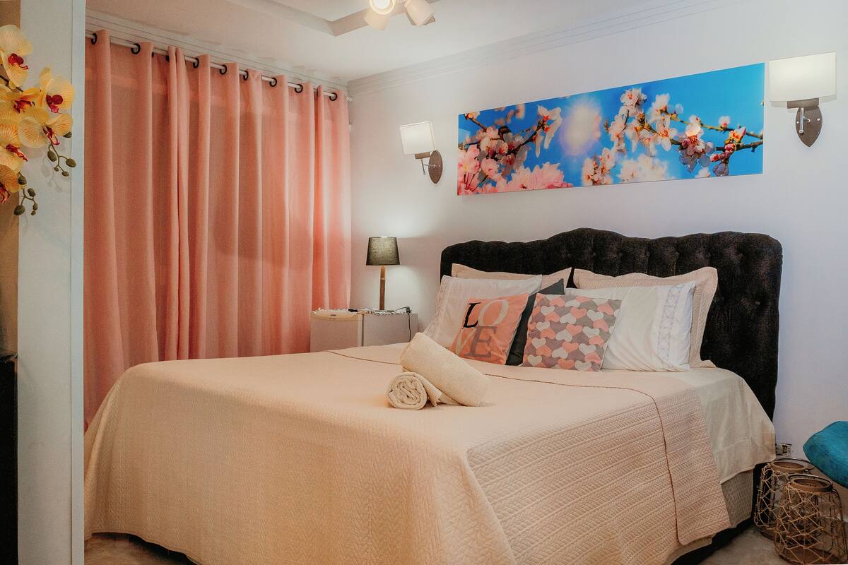 A bedroom with a bed, a nightstand, a lamp, a wall-mounted light, and a floral artwork.
