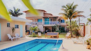 Une piscine entourée de palmiers, avec des chaises longues et un banc, et un bâtiment à deux étages doté de balcons.