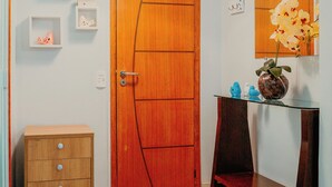 Un couloir avec une porte en bois, une petite armoire en bois, une console en verre surmontée d’un vase de fleurs et une étagère murale agrémentée d’objets décoratifs.