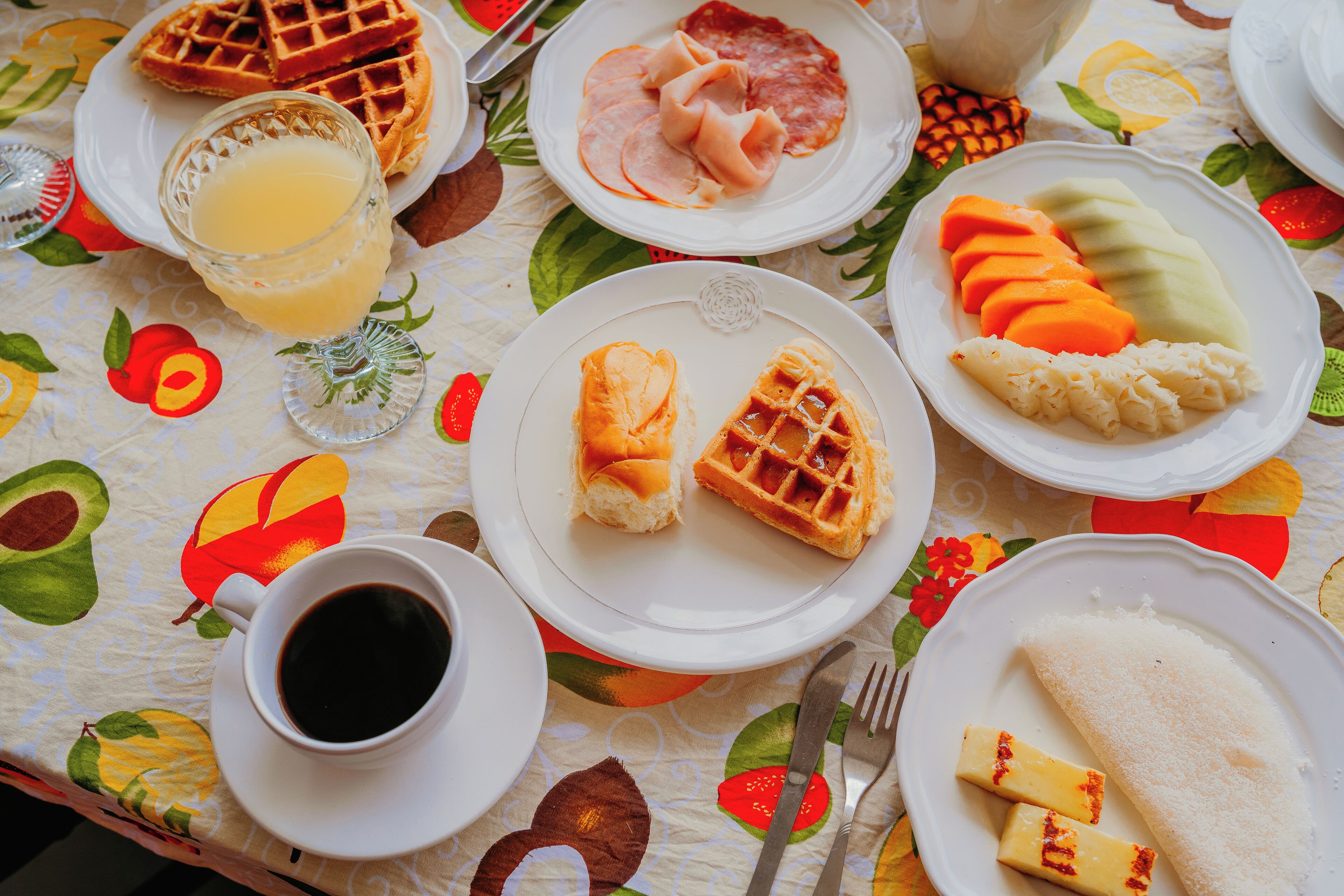 A breakfast table with waffles, ham, fruit, and a cup of coffee.