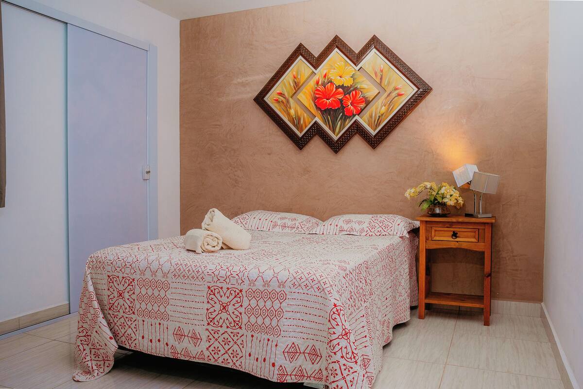 A neatly made bed with a patterned bedspread, a bedside table with a lamp, and a framed floral artwork on the wall.