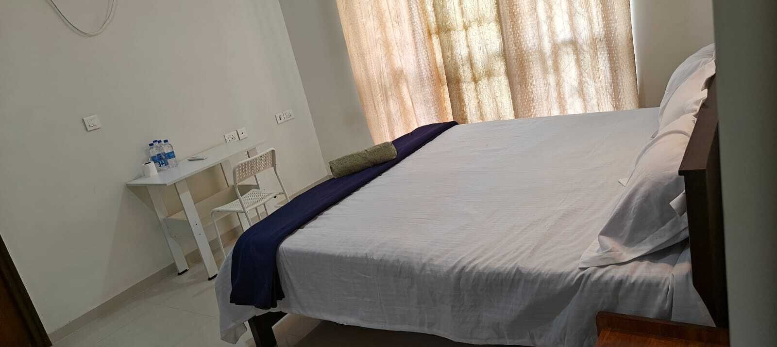 Une chambre à coucher avec un lit, une petite table blanche et une chaise, ainsi qu’une fenêtre avec des rideaux.
