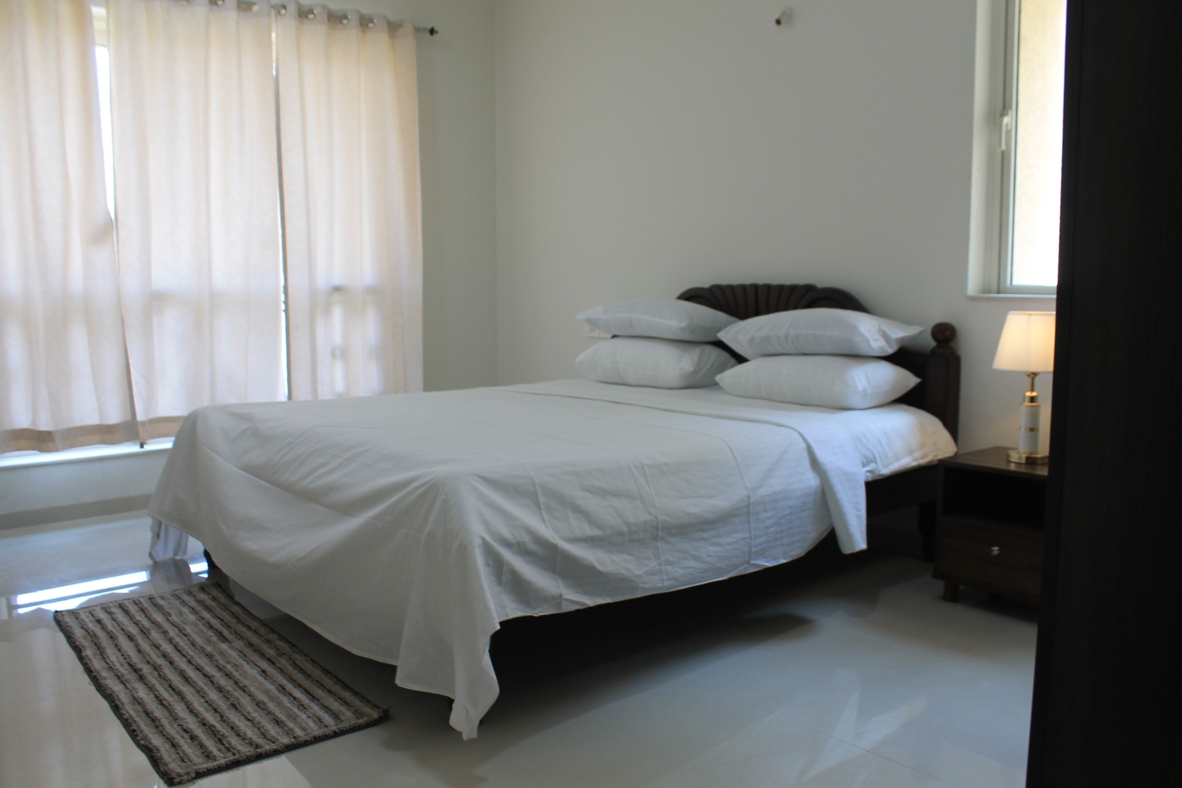 A neatly made bed with white linens, a bedside table with a lamp, and a window with curtains.