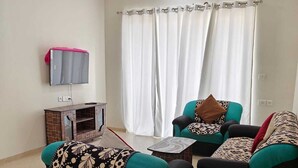 Un salon avec deux canapés noirs, dont l’un est agrémenté d’un plaid à motifs, une petite table basse, un téléviseur fixé au mur et un ventilateur de plafond.
