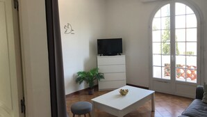 A modern living room with a white table, a grey sofa, a TV on a white cabinet, a potted plant, and a large arched window. - Superbe T2 en Centre Ville, à 400m du Port Climatisé Avec Parking Privé (Bandol)
