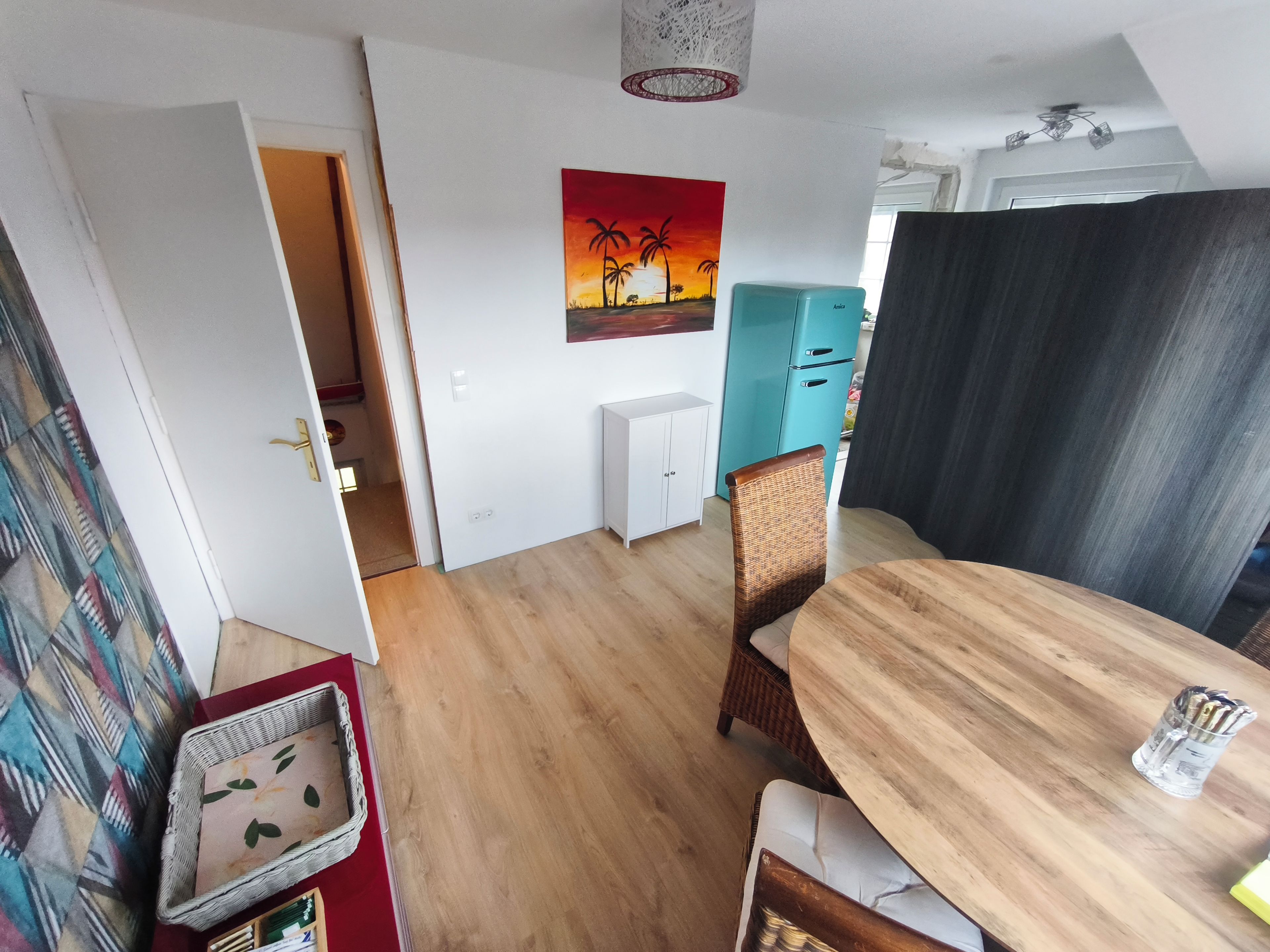 A dining area with a wooden table and chairs, a painting on the wall, and a kitchenette in the background.